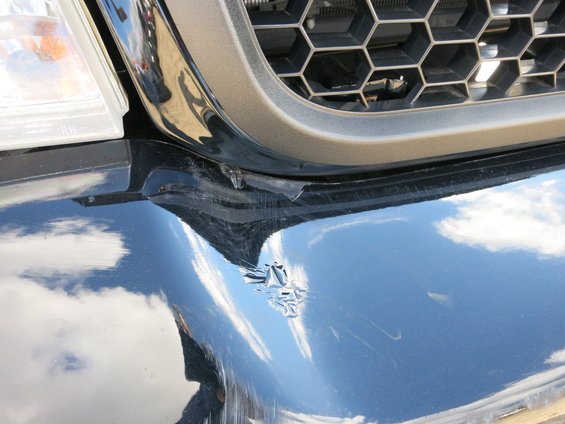Black car front bumper with damage and scratches, reflecting clouds and sky. Grille is visible above