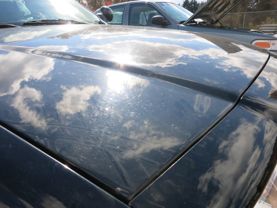 Dark blue car hood reflecting clouds, showing scratches and sun glare. Another car with its hood open is visible behind