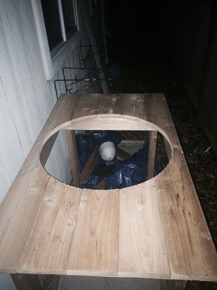 Unfinished wooden table with a large oval cutout, revealing black plastic bags and wooden supports underneath. The table is positioned next to a light-colored, paneled wall