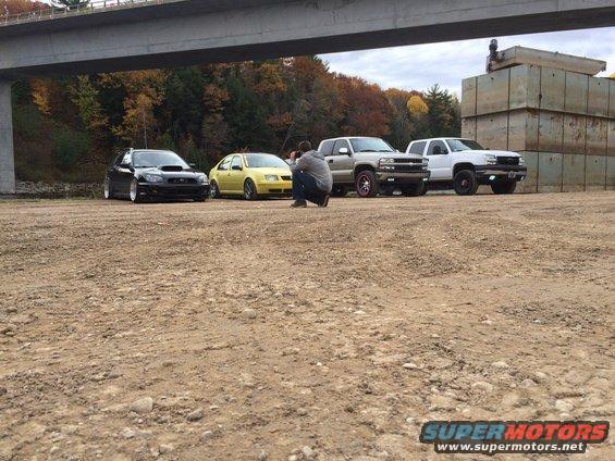 guy taking photo of four vehicles: black Subaru, yellow VW, silver truck, white truck