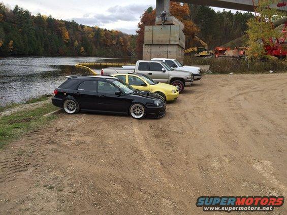 Black Subaru wagon, yellow sedan, and two pickup trucks parked on dirt near a river and bridge construction