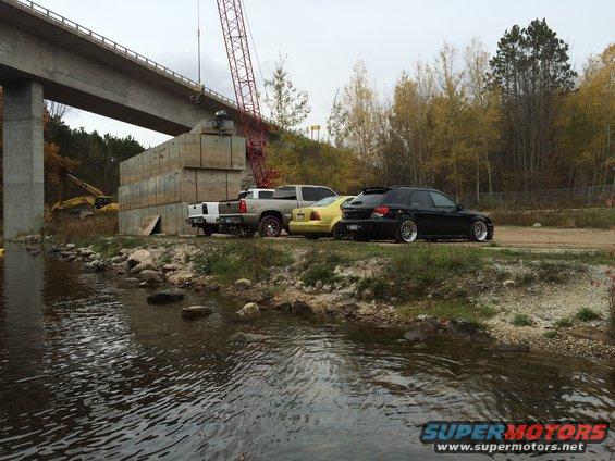 Construction site with a bridge, crane, concrete blocks, and several parked vehicles including a pickup truck, a yellow car, and a black hatchback