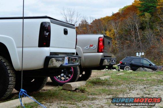 White pickup truck with pink rims, silver pickup truck with 4x4 badge, and black SUV parked on grassy terrain