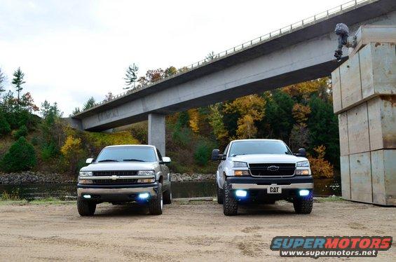 Two Chevrolet Silverado trucks parked near a bridge, one silver, one white. The white truck has a CAT license plate