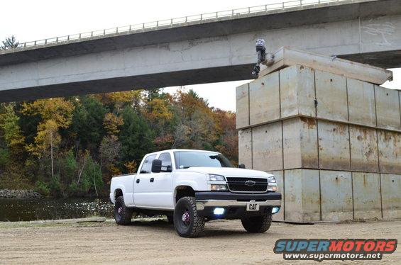 White pickup truck with aftermarket wheels and CAT badging on the grille, parked on dirt near a bridge and concrete blocks