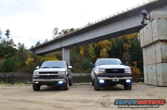 Two Chevrolet Silverado trucks parked on dirt, one silver, one white, under a concrete bridge