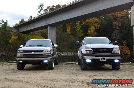 Two Chevrolet Silverado trucks parked under a bridge, one silver, one white. The white truck has a CAT license plate