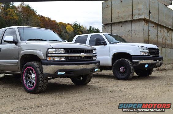Two Chevrolet Silverado trucks parked side-by-side, one silver with pink-accented wheels, the other white. Both are viewed from a 3/4 front angle