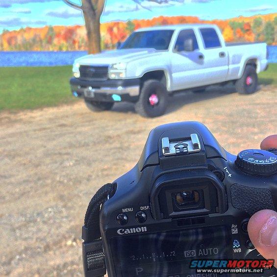 White 2006 Chevrolet Silverado 2500 pickup truck parked near a body of water with autumn foliage in the background. Camera in foreground