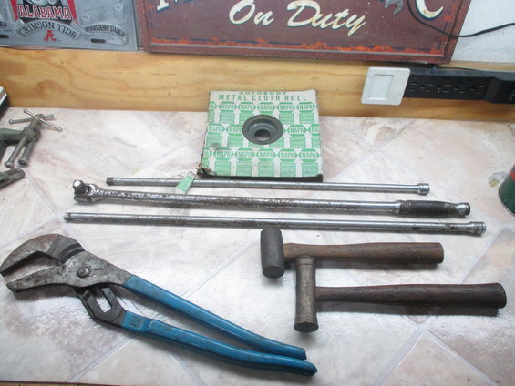 Pipe wrench, socket extensions, and hammers on a tiled surface. Box of NAPA autobody metal cloth roll in background