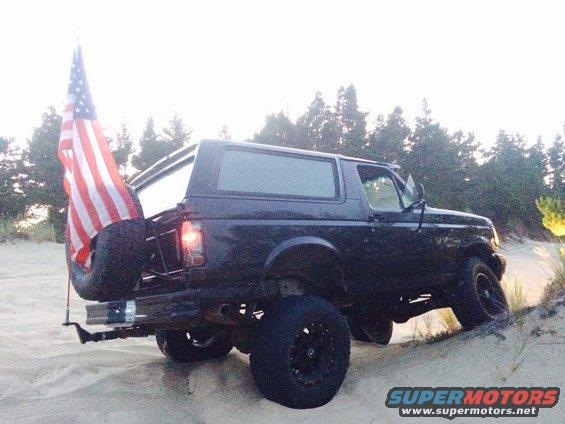 Black Ford Bronco with spare tire and American flag on rear bumper, driving on sand