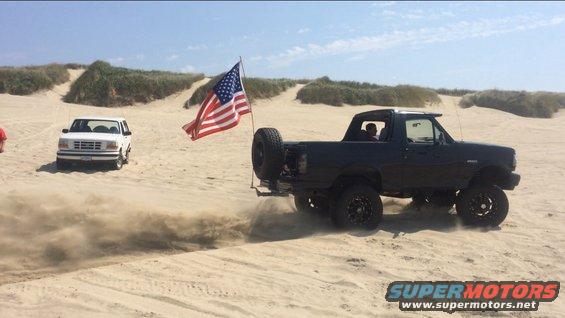 Black 1992 Ford Bronco with an American flag driving on sand dunes, lifted suspension, spare tire on rear