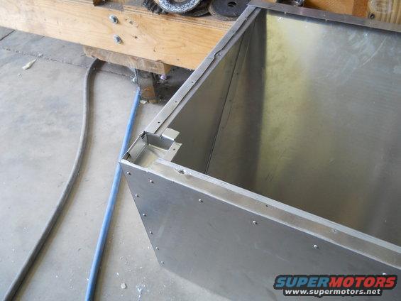 Metal box construction with visible rivets and seams, on a concrete floor next to a wooden workbench