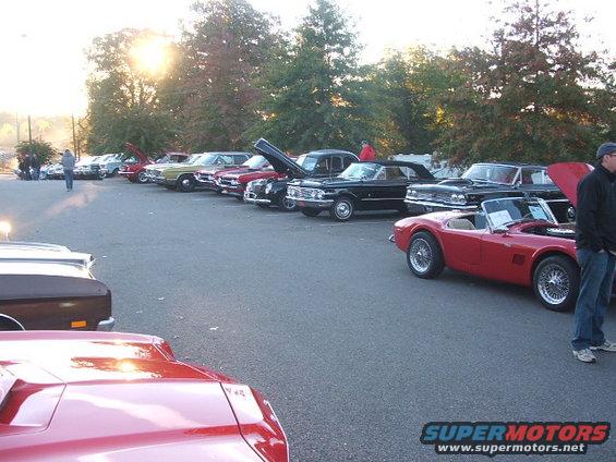 Several classic cars parked in a lot, including a red convertible sports car and a black convertible. Sun is setting