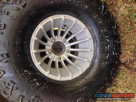 Close-up of a large off-road tire with a silver multi-spoke wheel. Grass is visible around the tire