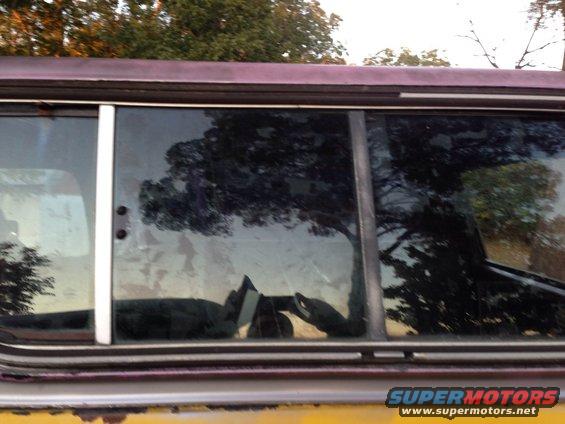 Yellow and maroon vehicle side window with reflections of trees and sky. A Supermotors logo is visible