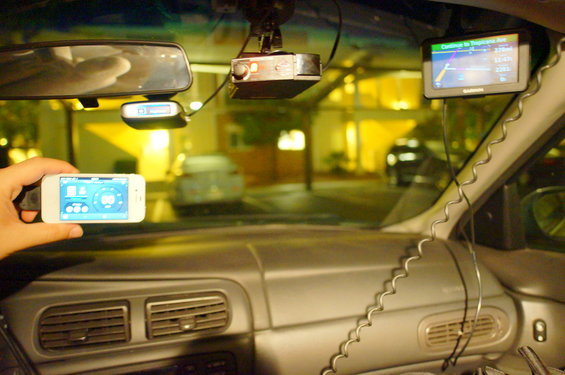 Hand holding phone showing app, dash cam, and GPS unit mounted on windshield