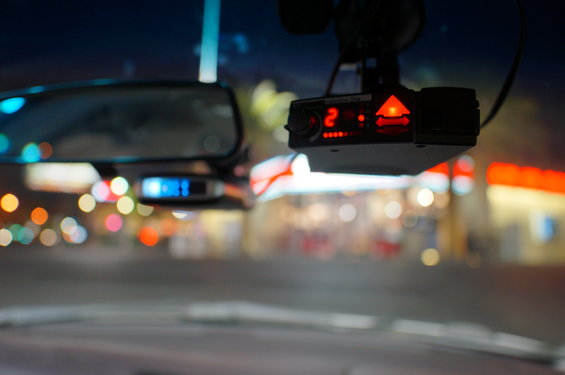 Radar detector with red lights and digital display, mounted on windshield near rearview mirror. Blurred lights in background