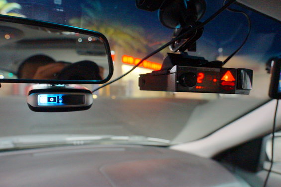 Radar detector with red lights and a dashcam mounted on a windshield, with a rearview mirror reflecting a person