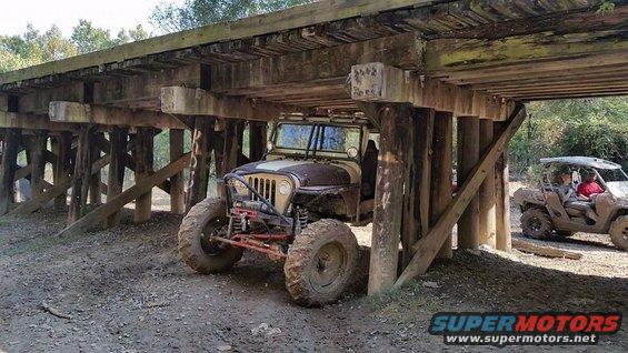 Modified Jeep parked under a wooden bridge, another UTV visible to the right