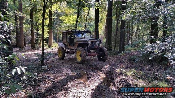 Modified Jeep CJ-7 with large tires and roll cage on a muddy forest trail