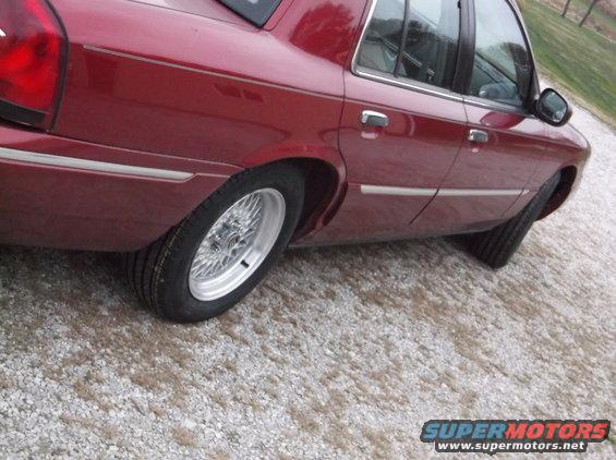 Red sedan rear quarter panel with chrome trim and wire wheels. Gravel surface