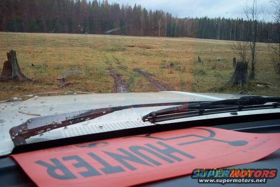View from inside a vehicle looking out at a muddy field and forest. Red paper with black text 'HUNTER' is on the dashboard