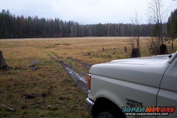 White 1990 Ford Bronco parked in a muddy field with trees in the background