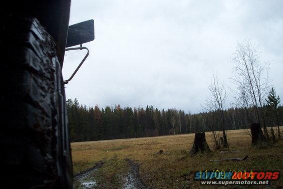 Close-up of a muddy tire and fender with a field and forest in the background