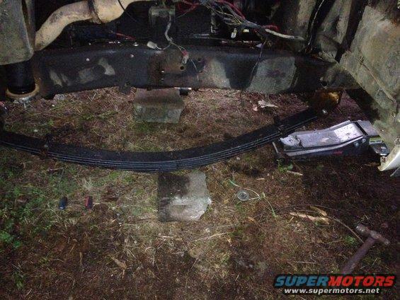 Truck frame with leaf spring removed, supported by blocks and a jack. Wires and exhaust pipe visible