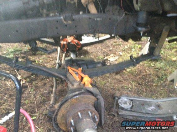 Truck chassis underside with leaf springs, brake rotor, and orange straps. Supermotors.net logo visible