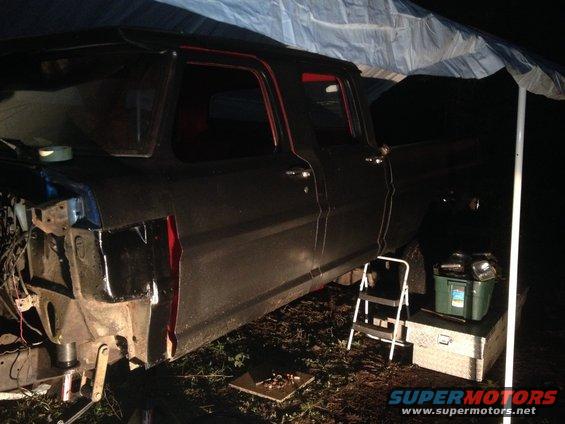 Side view of a dark-colored pickup truck, possibly undergoing repairs, parked under a tarp. A step ladder and toolboxes are nearby