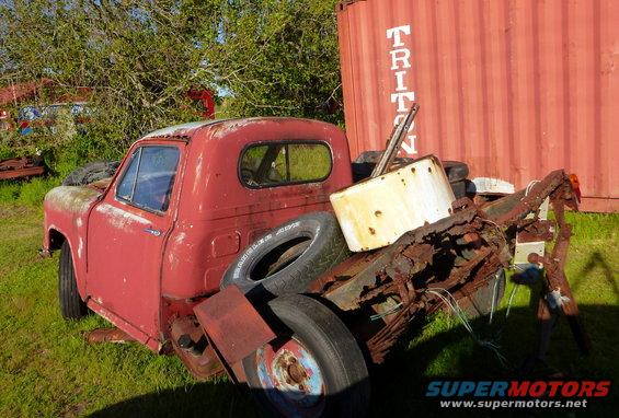 Red pickup truck with rust and damage, loaded with tires and scrap metal, parked outdoors next to a red shipping container