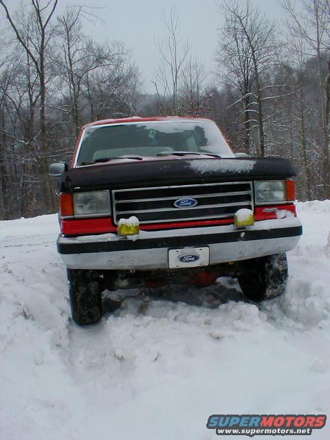 Front view of a red 1990 Ford Bronco in deep snow, with trees in the background
