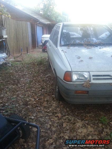 White hatchback parked in a yard with leaves on the ground. Another vehicle is visible in the background