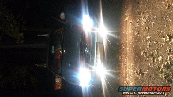 2000 Ford Crown Victoria, front view, headlights on, dark exterior, parked on dirt road with leaves