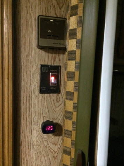 RV interior panel with a thermostat, a start/stop switch, and a digital voltage meter showing 12.5