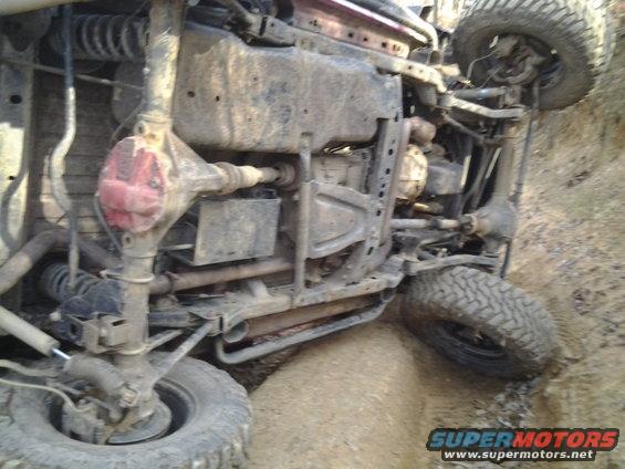 Off-road vehicle flipped onto its side in dirt, showing undercarriage and suspension. Supermotors.net logo visible