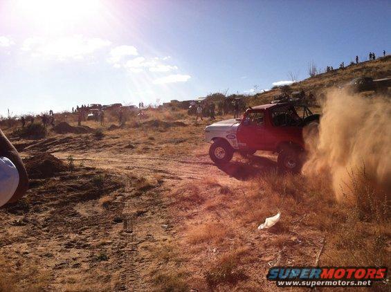 Red and white Ford Bronco kicking up dust on an off-road course with spectators on a hill in the background