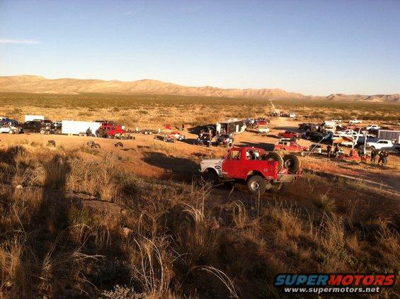 Red off-road truck with spare tire on back, driving on dirt path. Many other vehicles and trailers in background desert scene