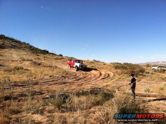 Red and white Ford Bronco driving on a dusty, dry hill with tire tracks. A person stands nearby with a yellow rope
