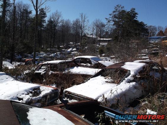 Junkyard scene with several rusty cars covered in snow; one car shows a grille and headlight area
