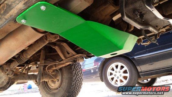 Underside view of a vehicle's frame with a green skid plate installed. Another car is visible in the background