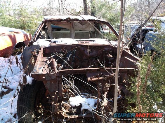 Rusted out car front end in a junkyard with snow on the ground. Another car is visible to the left