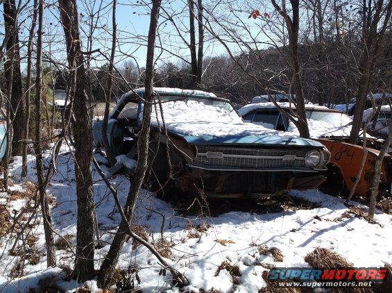 Snow-covered blue classic car in a wooded junkyard.