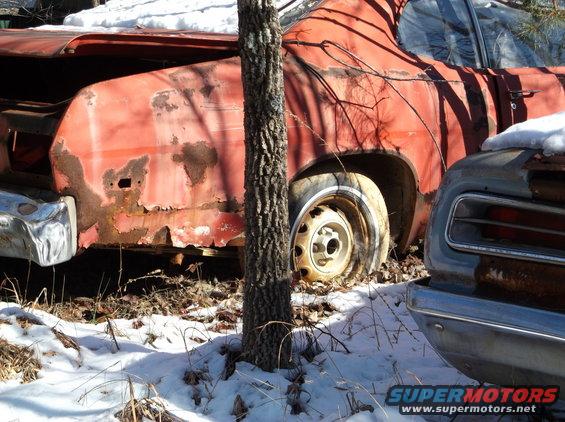 Rusty red vintage car rear quarter panel and wheel, with another car's front end in foreground, snow on ground.