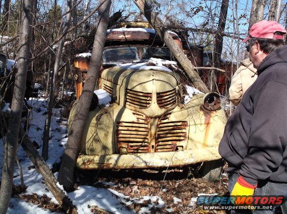 Rusty vintage yellow truck in a wooded area with snow, viewed from the front.