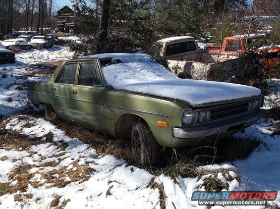 Green sedan covered in snow, parked in a junkyard with other vehicles