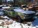 1974 Dodge Challenger - A green sedan sits in a snowy junkyard, surrounded by trees and other abandoned vehicles.
