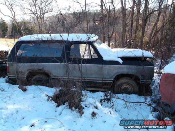 Side view of a weathered, two-tone black and grey SUV covered in snow and surrounded by trees.